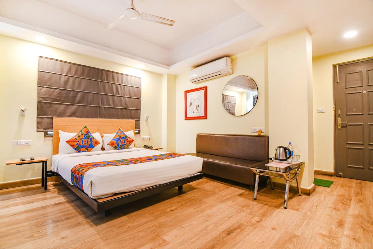 A hotel room with a bed, a sofa, a small table with a kettle, a mirror, and an air conditioning unit.