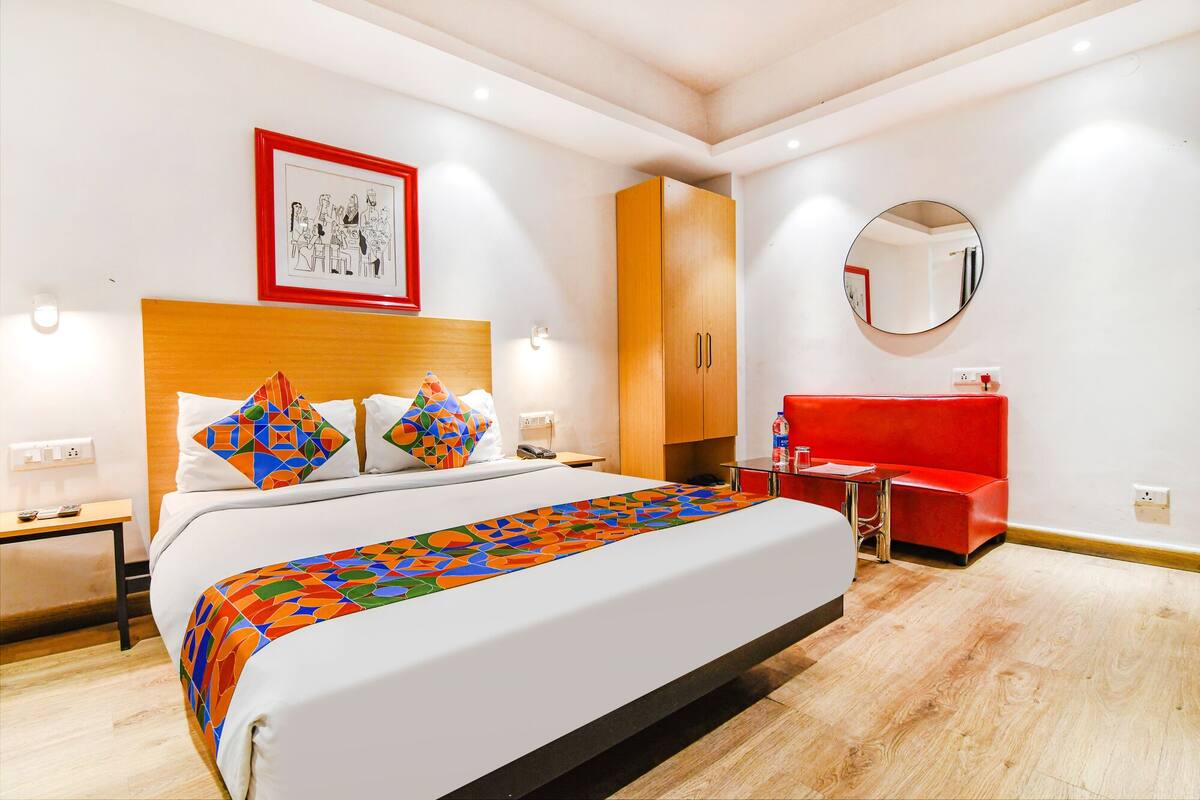 A hotel room with a large bed, a red vanity, a wooden wardrobe, a framed picture on the wall, and a round mirror.