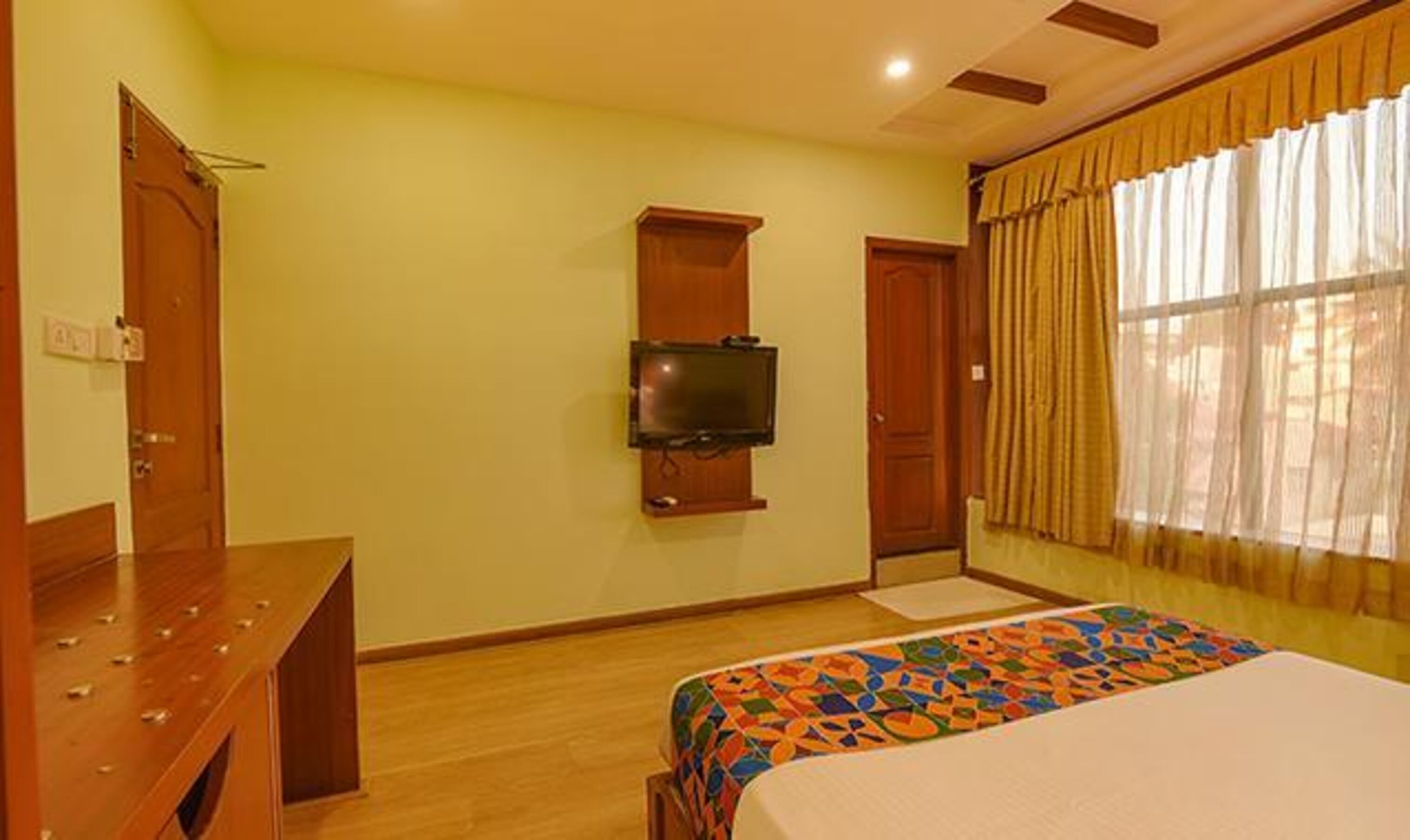 A hotel room with a wooden bed, a television mounted on the wall, and a window with curtains.