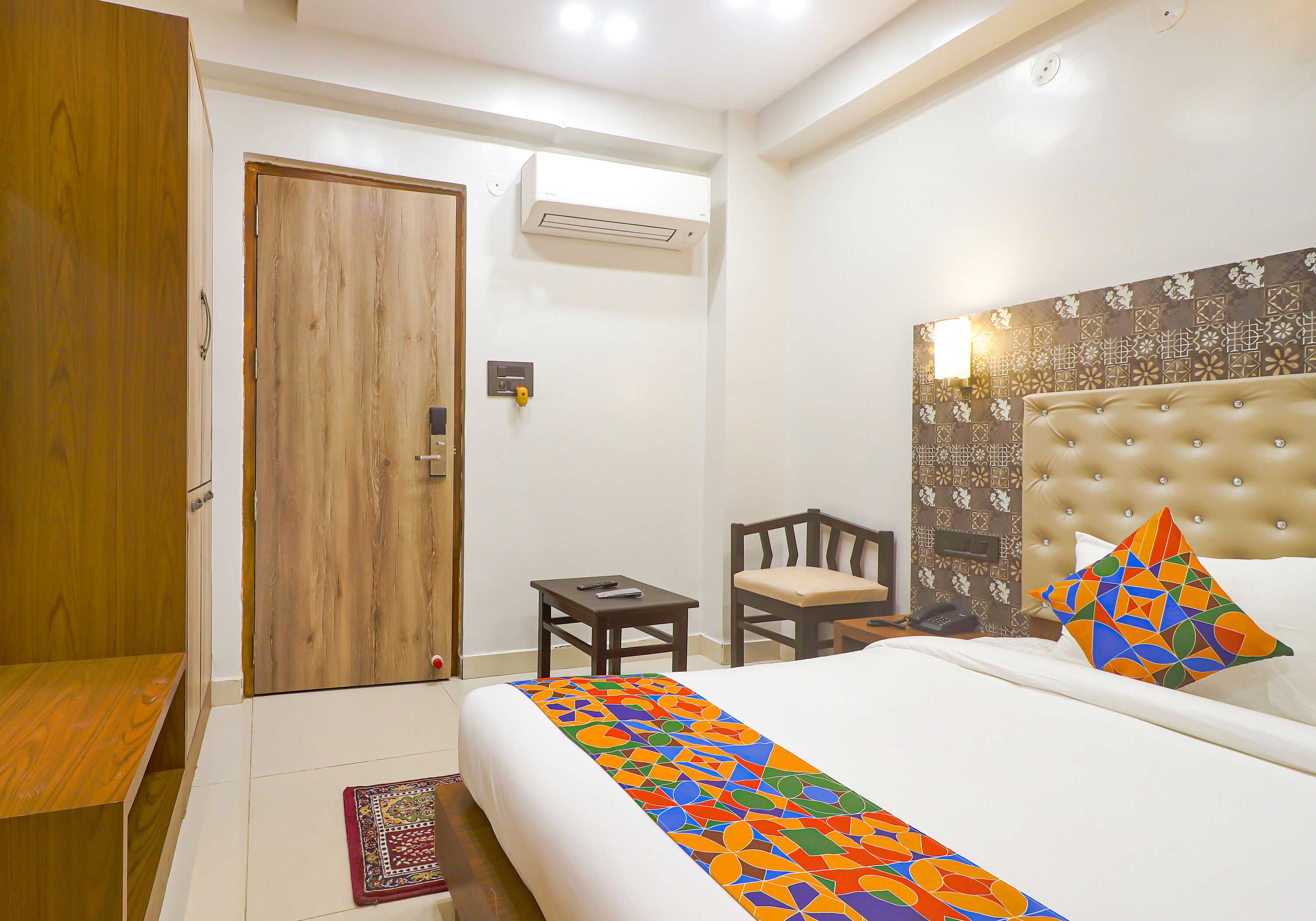 A hotel room with a bed, a small table, a chair, a wardrobe, and an air conditioning unit.