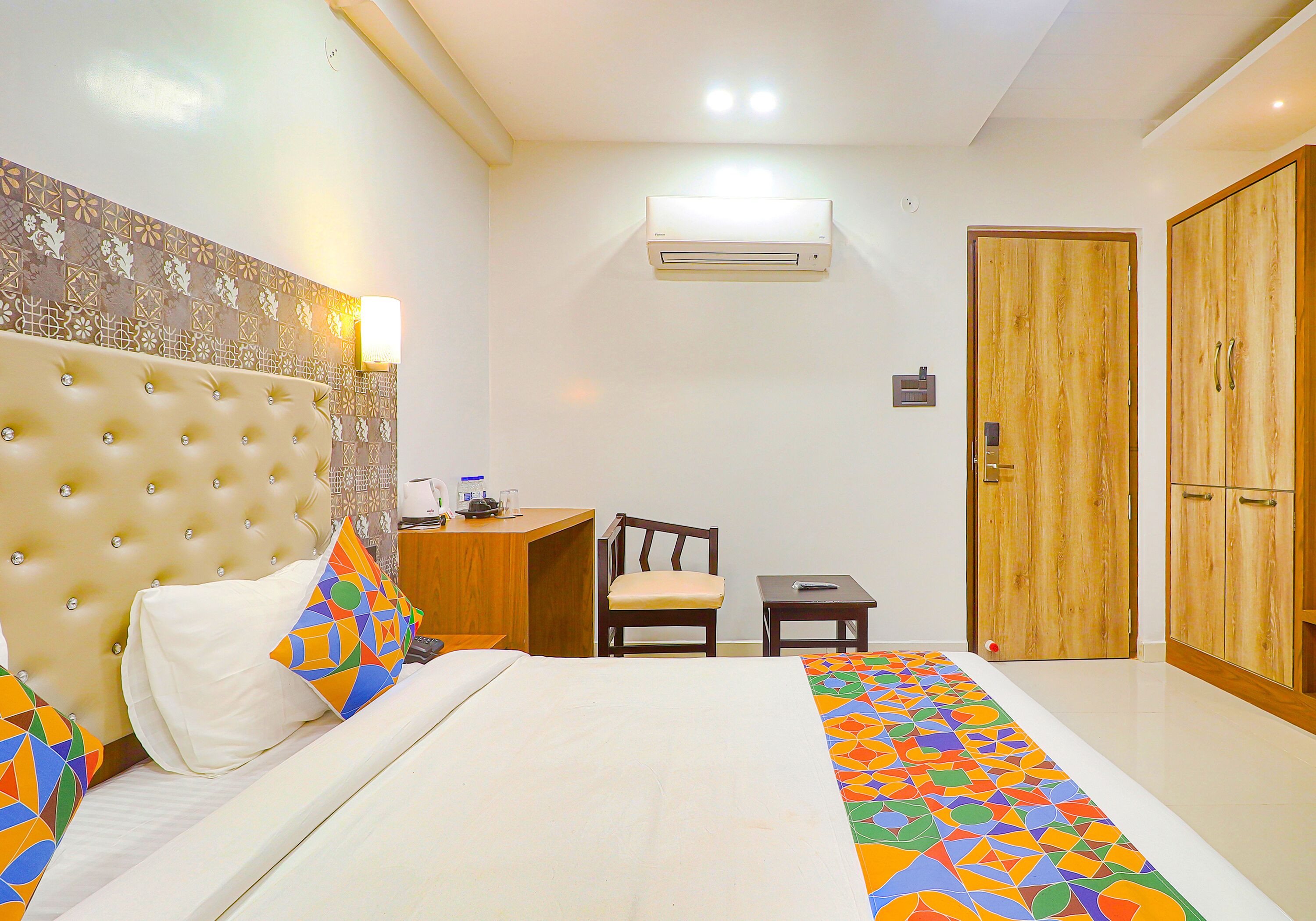 A hotel room with a tufted headboard, a bed with colorful geometric bedding, a wooden desk with a chair, a small table, and a wardrobe with two doors.