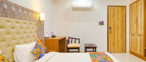 A hotel room with a tufted headboard, a bed with colorful geometric bedding, a wooden desk with a chair, a small table, and a wardrobe with two doors.