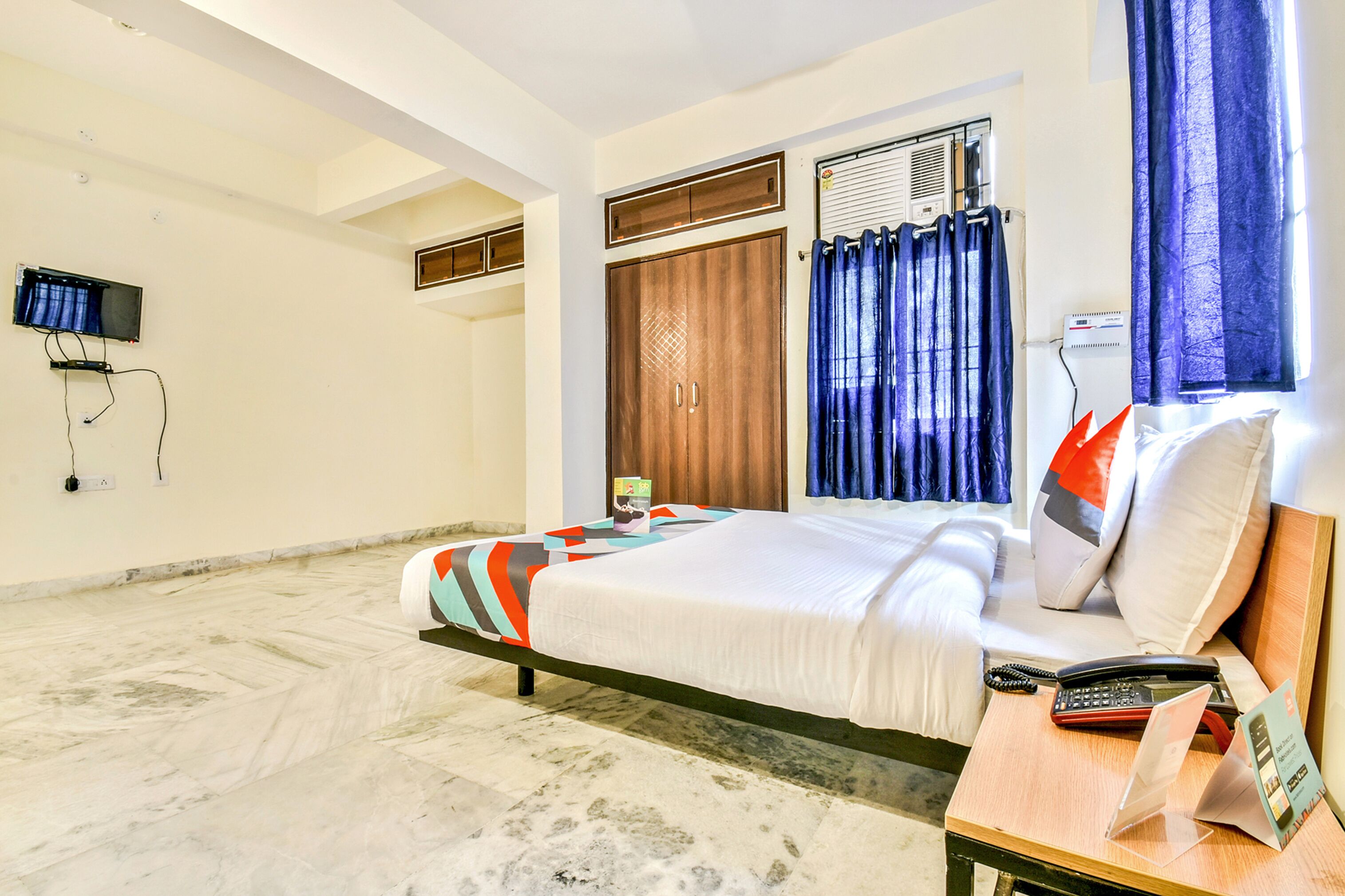 A hotel room with a bed, a desk with a phone and a brochure, a TV mounted on the wall, and a window with curtains.