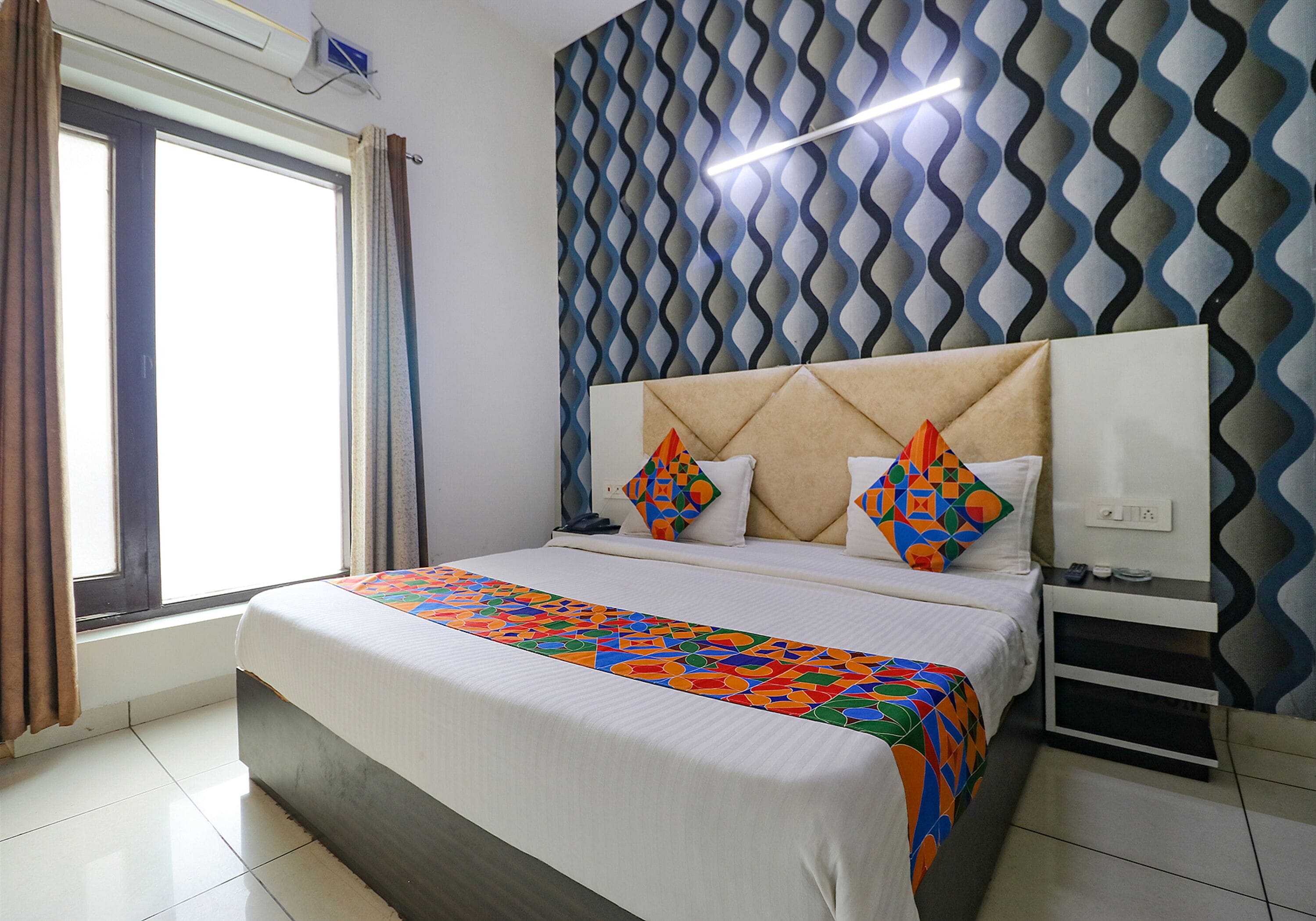 A neatly made bed with a colorful bedspread, a headboard with geometric patterns, a bedside table with a lamp, and a window with curtains.