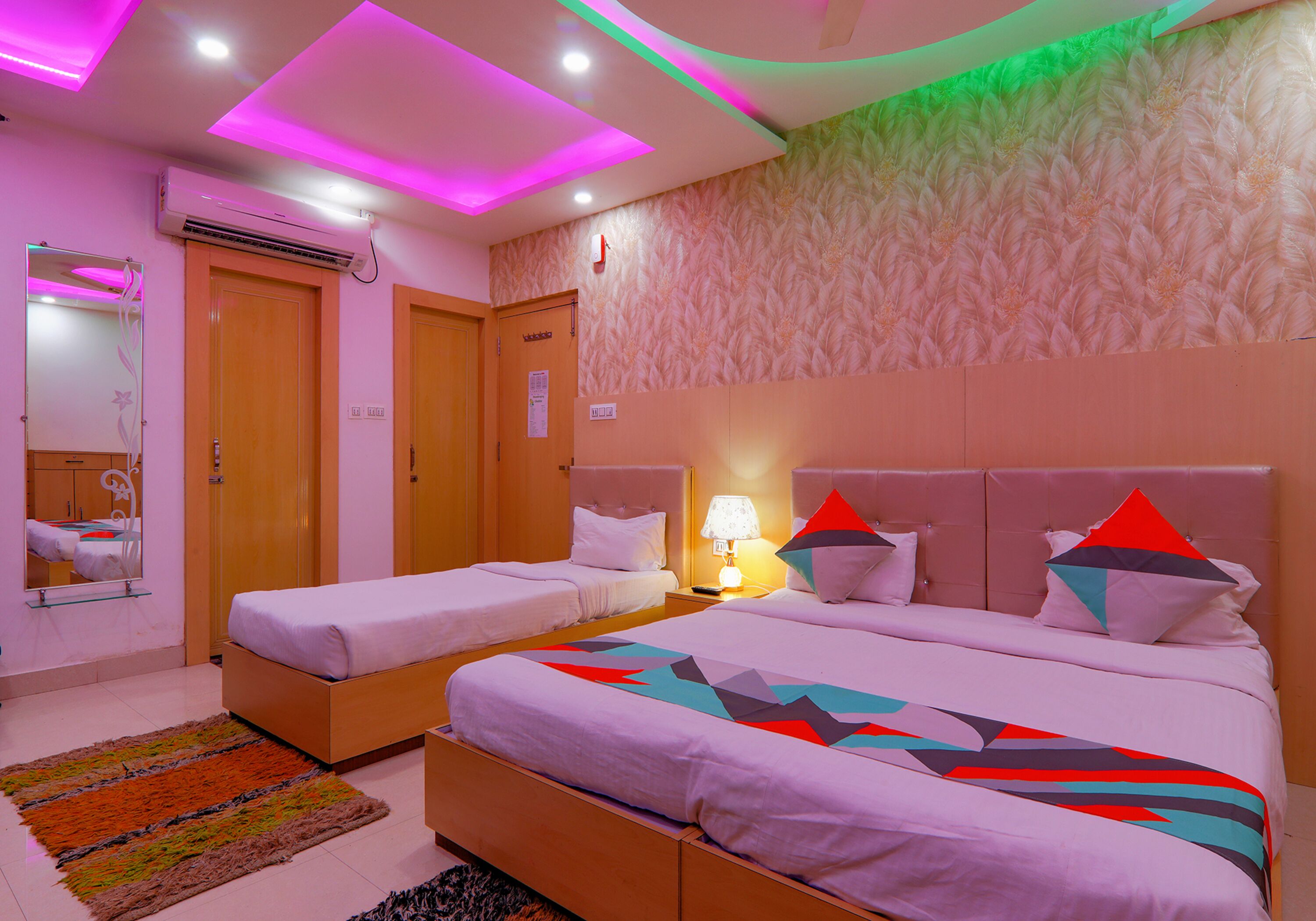 Habitación de hotel con dos camas individuales, una cama grande, un espejo, una mesita de noche con lámpara y un papel tapiz estampado.