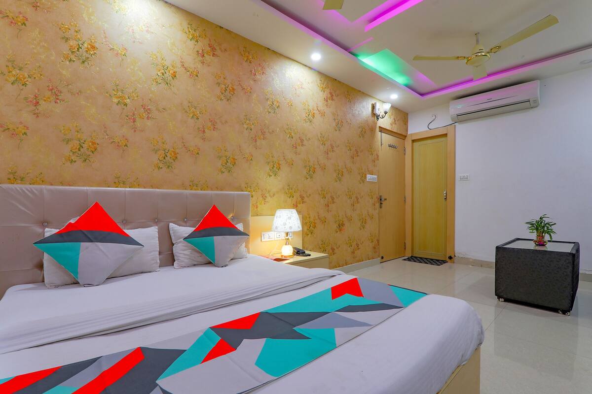 A hotel room with a bed, a nightstand, an air conditioning unit, and a small table with a plant.