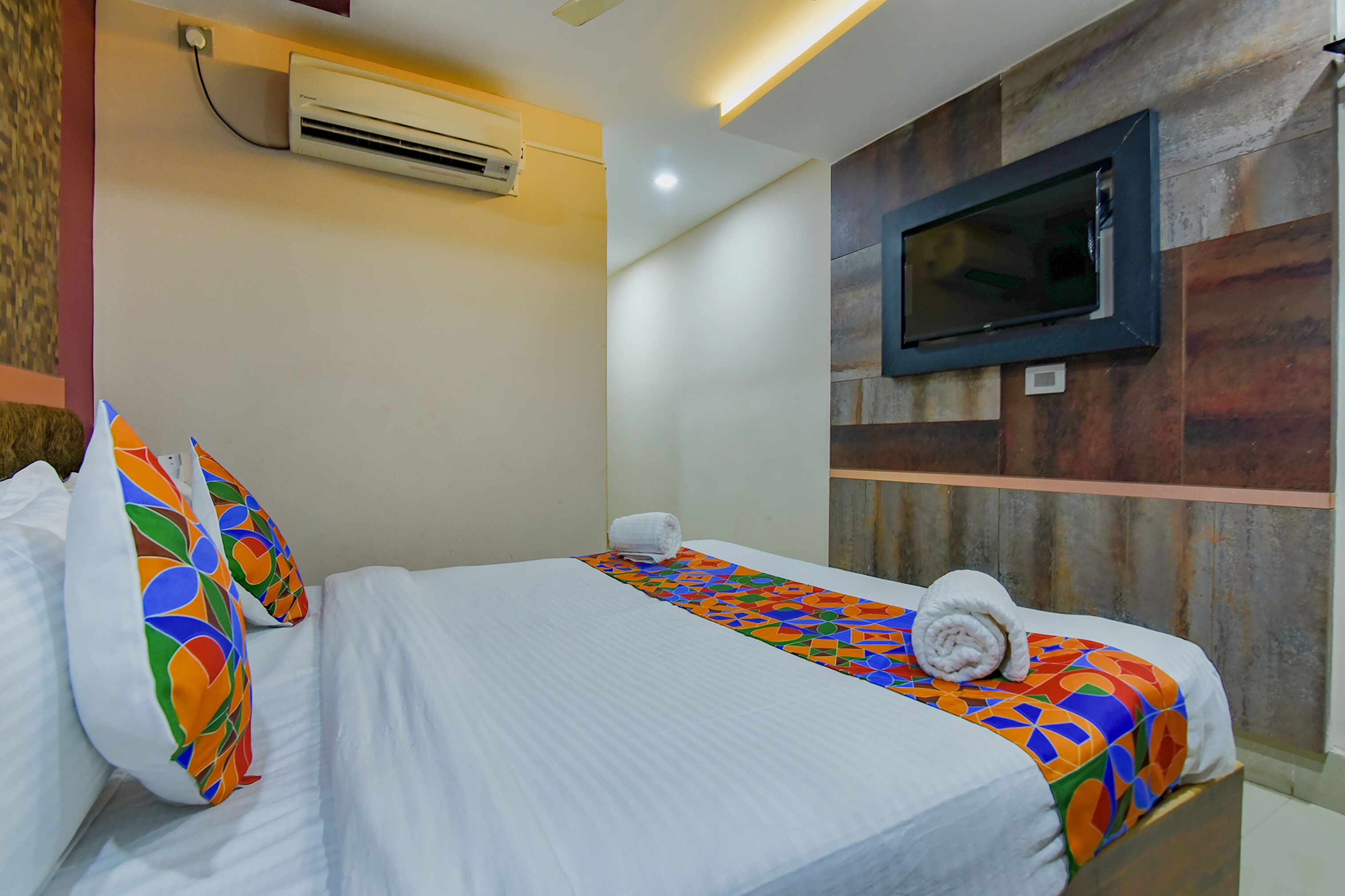 A hotel room with a bed, colorful pillows, a TV mounted on the wall, and an air conditioning unit.