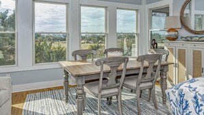 A dining area with a wooden table and chairs, large windows offering a view of greenery, and a mirror on the wall. - Golf course views on Fort Holmes Trail overlooking hole number 10! Upper deck taking in island views (Bald Head Island)