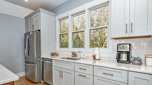 A modern kitchen with white cabinetry, a stainless steel refrigerator, a coffee maker, and a window with a view of trees. - Golf course views on Fort Holmes Trail overlooking hole number 10! Upper deck taking in island views (Bald Head Island)