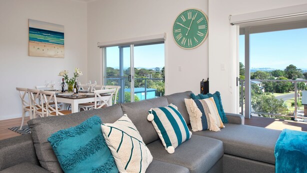 A modern living room with a grey sofa, teal and white pillows, a dining area with a white table and chairs, and a large green clock on the wall.