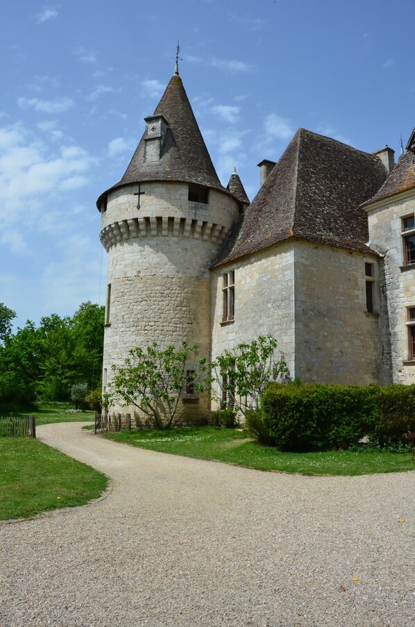 A stone castle with a round tower and a conical roof, surrounded by a gravel path and greenery.