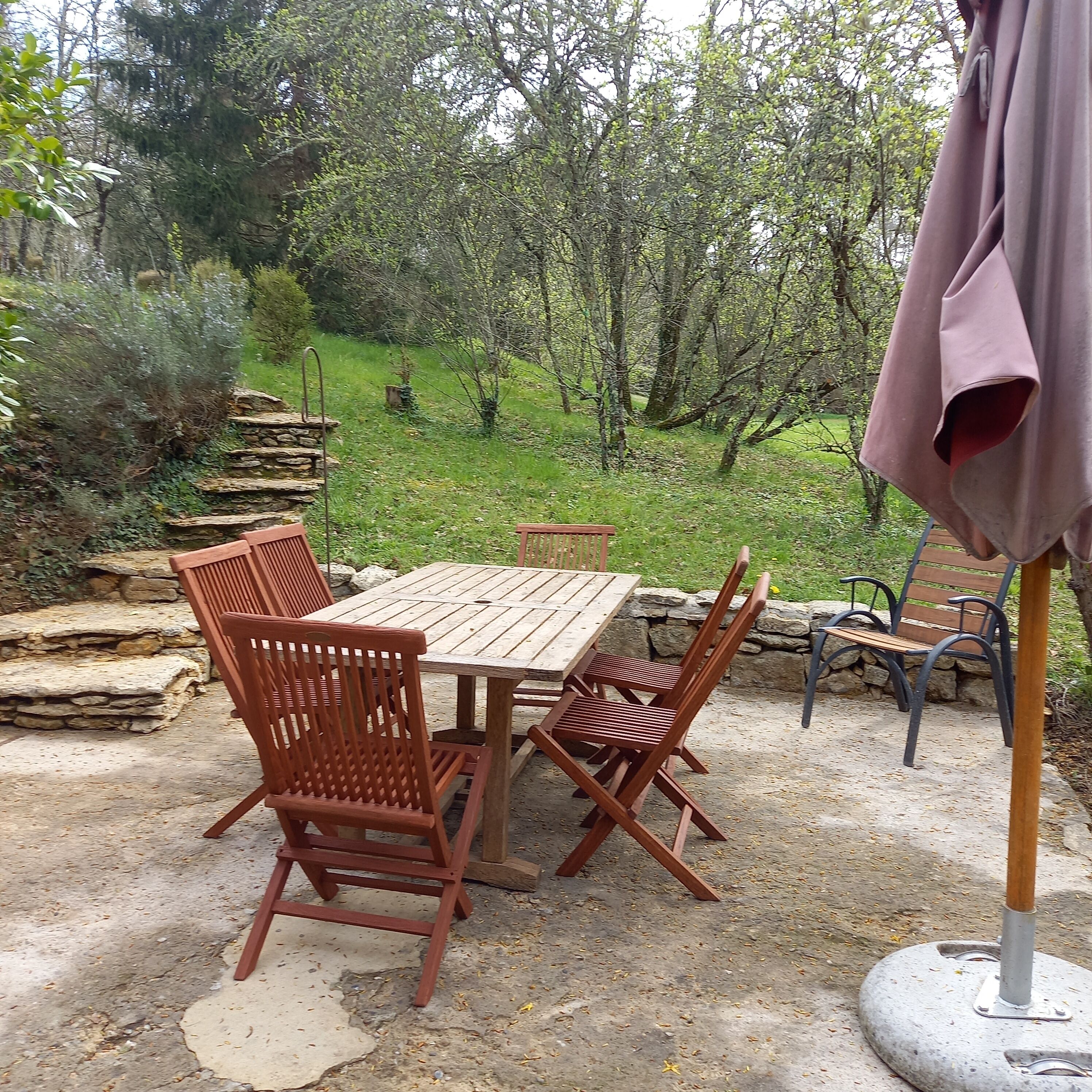 An outdoor patio with a wooden table and chairs, surrounded by stone walls and greenery.