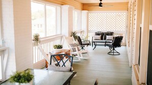A covered porch with wooden flooring, white railings, and ceiling fans. There are several chairs and a table with cushions.