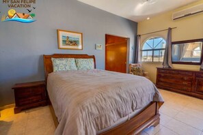 Une chambre à coucher avec un grand lit, une table de chevet en bois, une commode, un tableau encadré au mur et une fenêtre avec des rideaux.