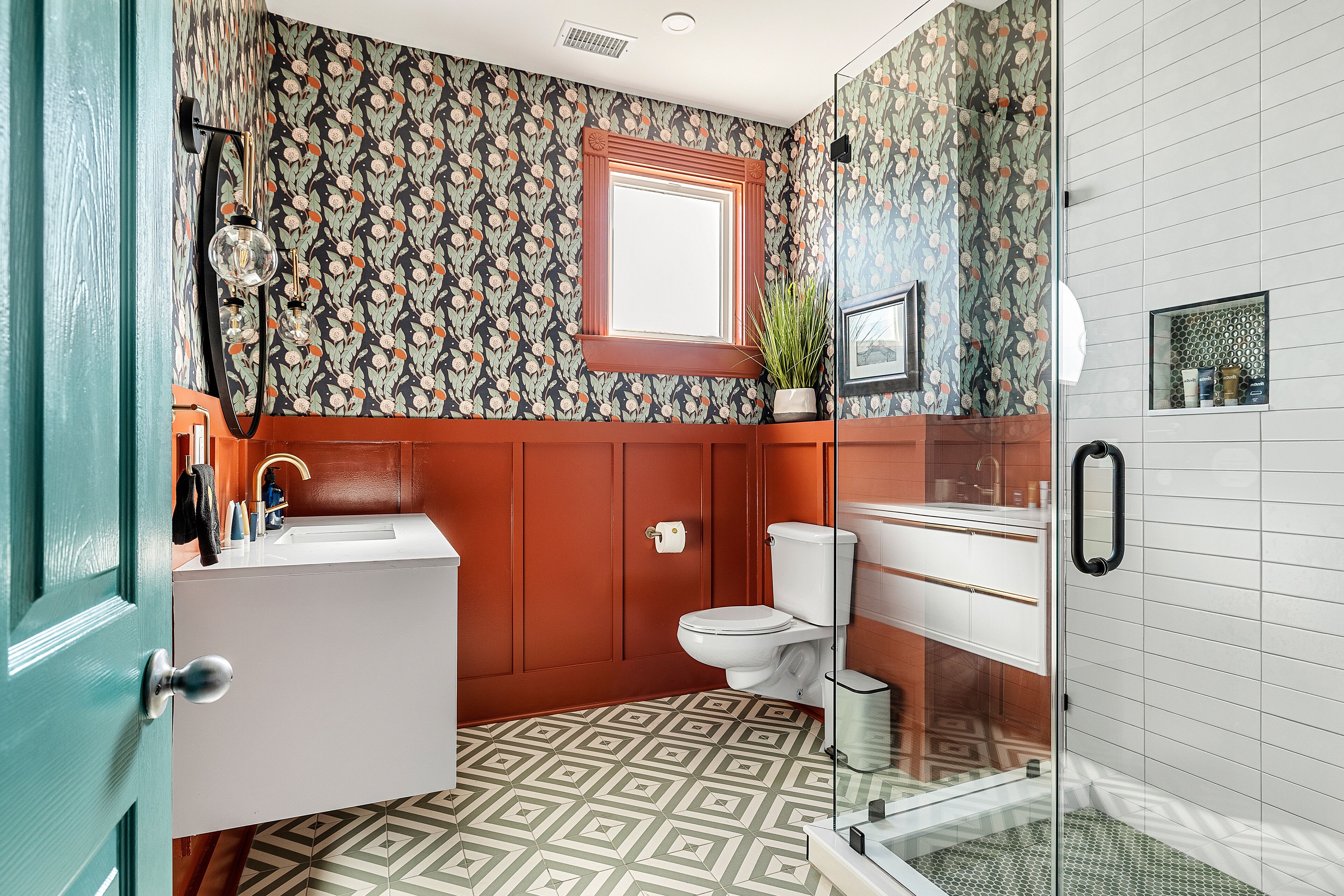 A bathroom with a patterned wallpaper, a window, a toilet, a sink, and a shower area.