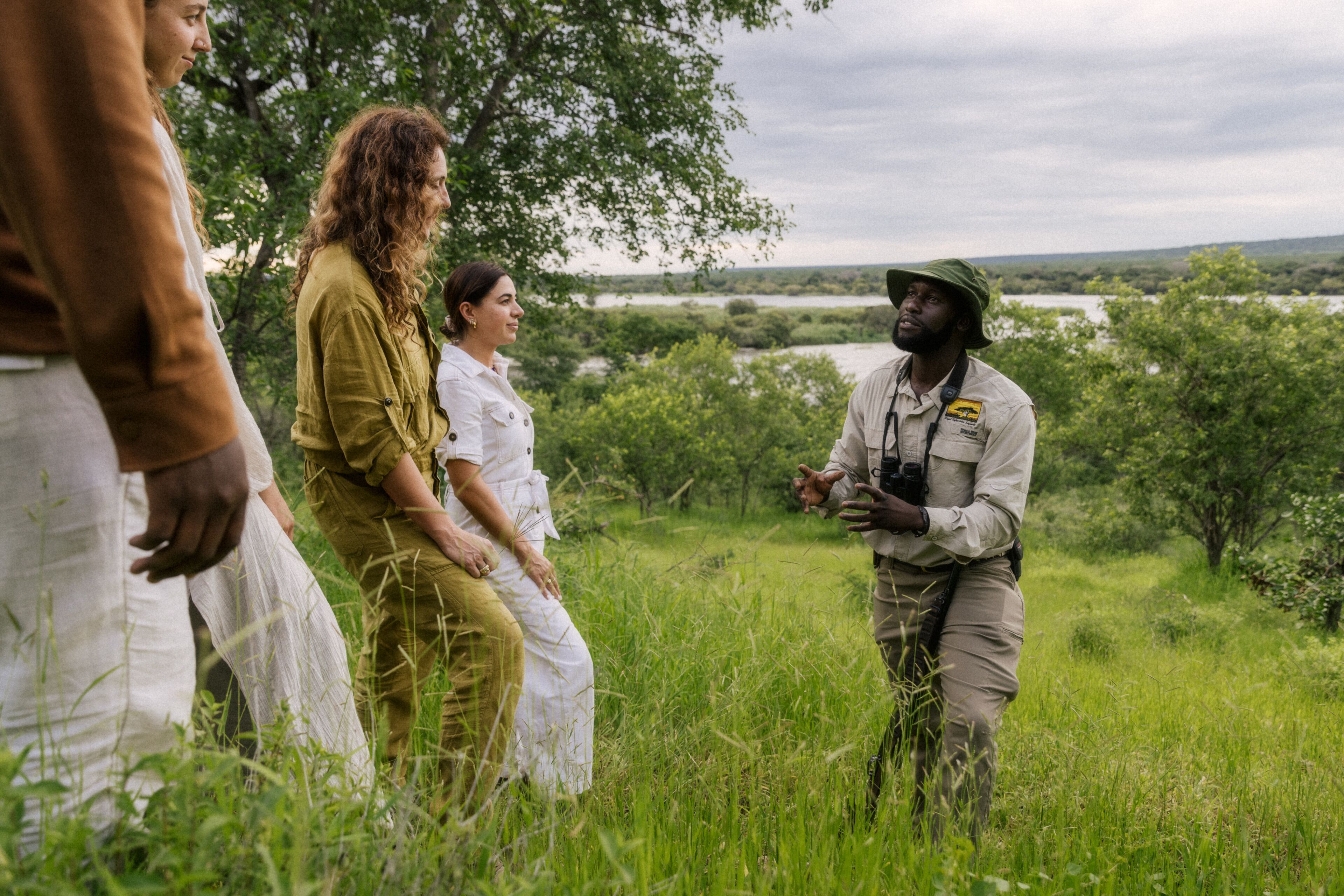 A group of people in a grassy outdoor area with trees and a body of water in the background.