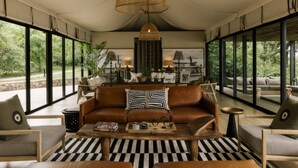 A spacious living area with a brown leather sofa, black and white striped rug, and large windows offering a view of greenery outside. - Zambezi Sands By Batoka (Victoria Falls)
