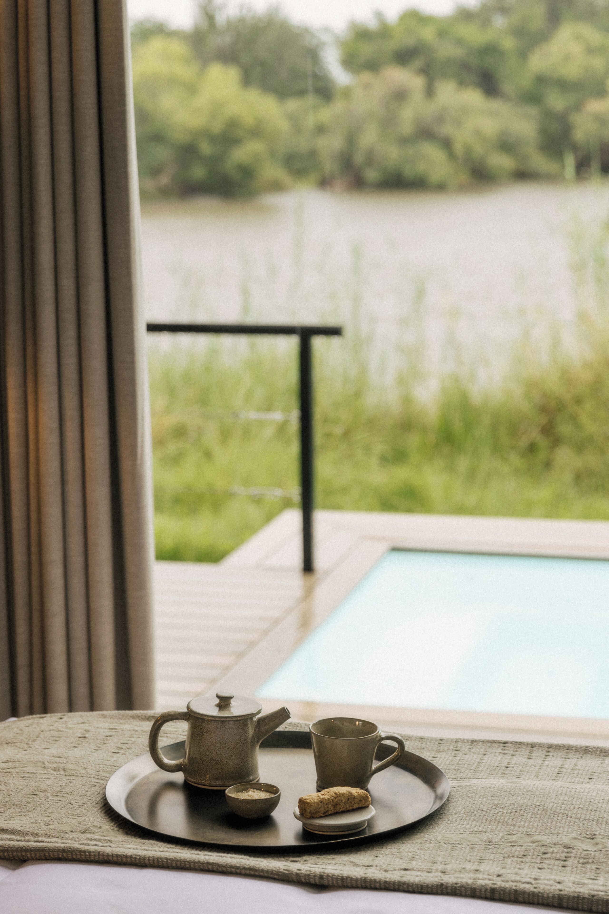 A table with a teapot, cup, and biscuit by a window overlooking a body of water.