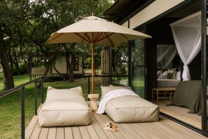 A deck with two beige pillows, a wooden table, and a large umbrella. - Zambezi Sands By Batoka (Victoria Falls)