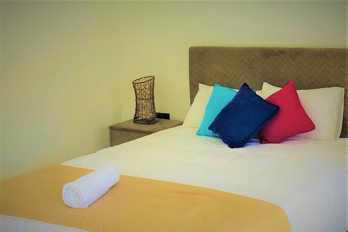 A neatly made bed with a rolled white towel, a headboard with a herringbone pattern, and multiple pillows in various colors.