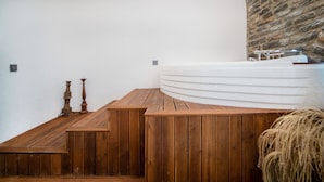 Eine Holzterrasse führt zu einer weißen Badewanne in einem Spa-Bereich mit einer Steinwand und einem kleinen Tisch mit einer Kerze.