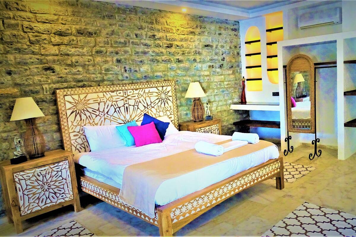 A bedroom with a stone wall, a wooden bed frame, a nightstand, a lamp, a mirror, and a shelf with decorative items.