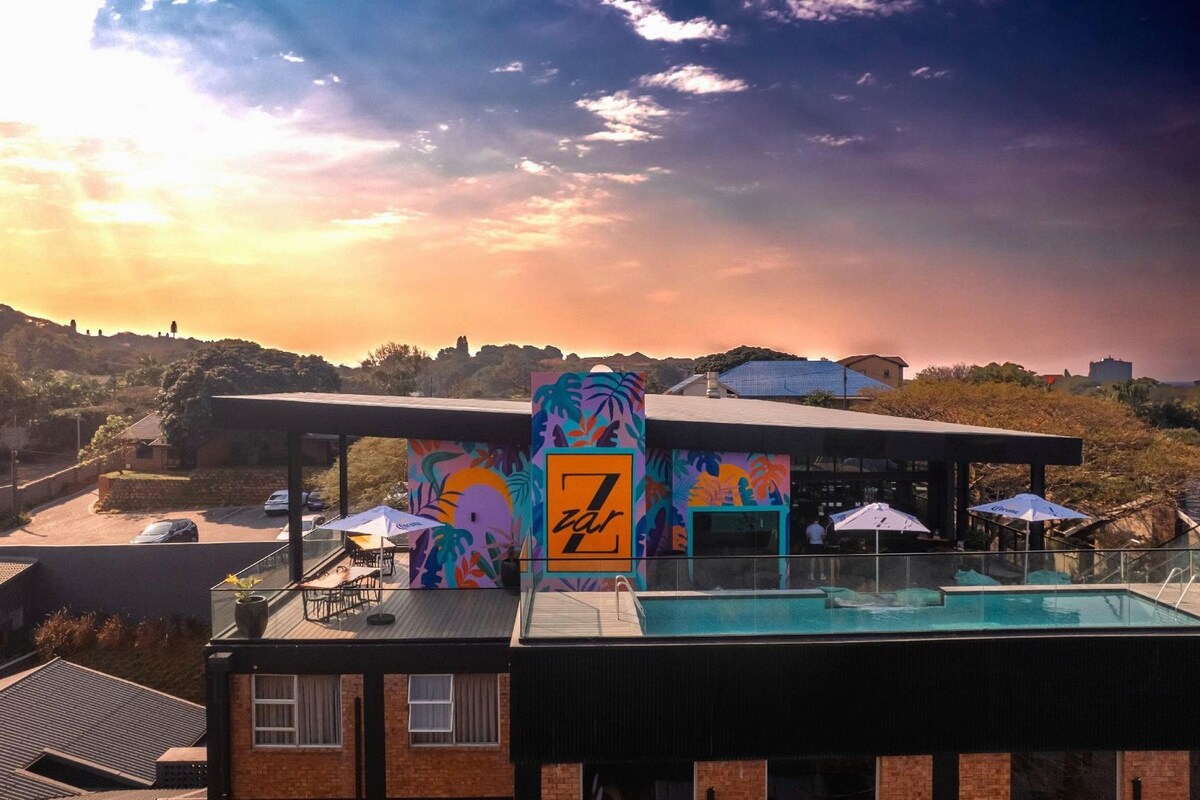 A rooftop pool area with a view of the surrounding landscape, a covered seating area, and a building with a colorful mural.