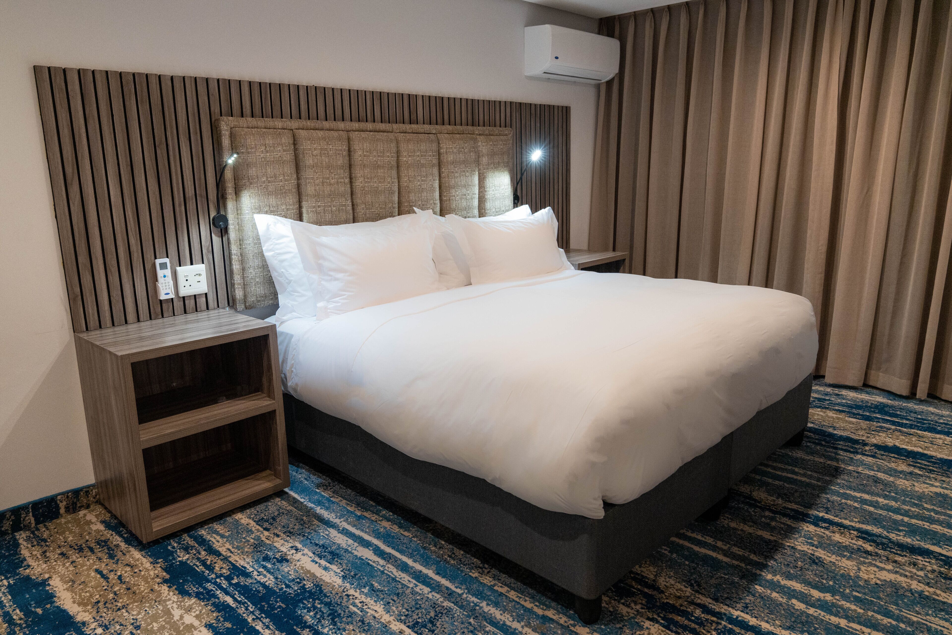 A hotel room with a large bed, a wooden headboard, a nightstand, and a blue striped carpet.