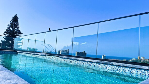 A clear blue pool with a tiled edge, a glass railing, and a view of the ocean and buildings in the background. - Zar Hotel (Kingsburgh)