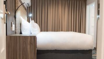Uma cama bem arrumada com roupa de cama branca, uma mesa de cabeceira de madeira e uma luminária fixada na parede.