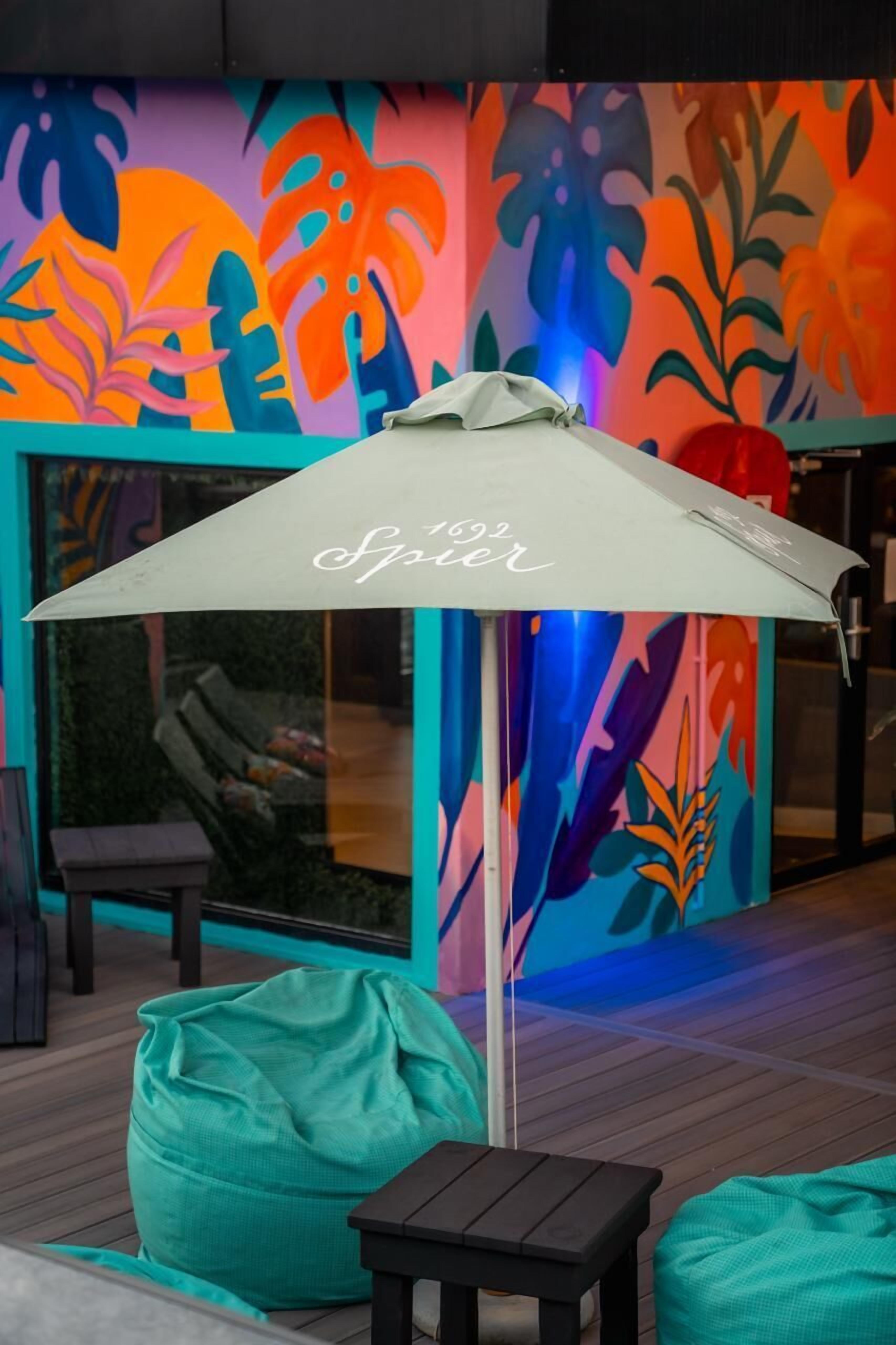Un área de estar moderna al aire libre con un mural colorido, un paraguas blanco y puffs color turquesa.