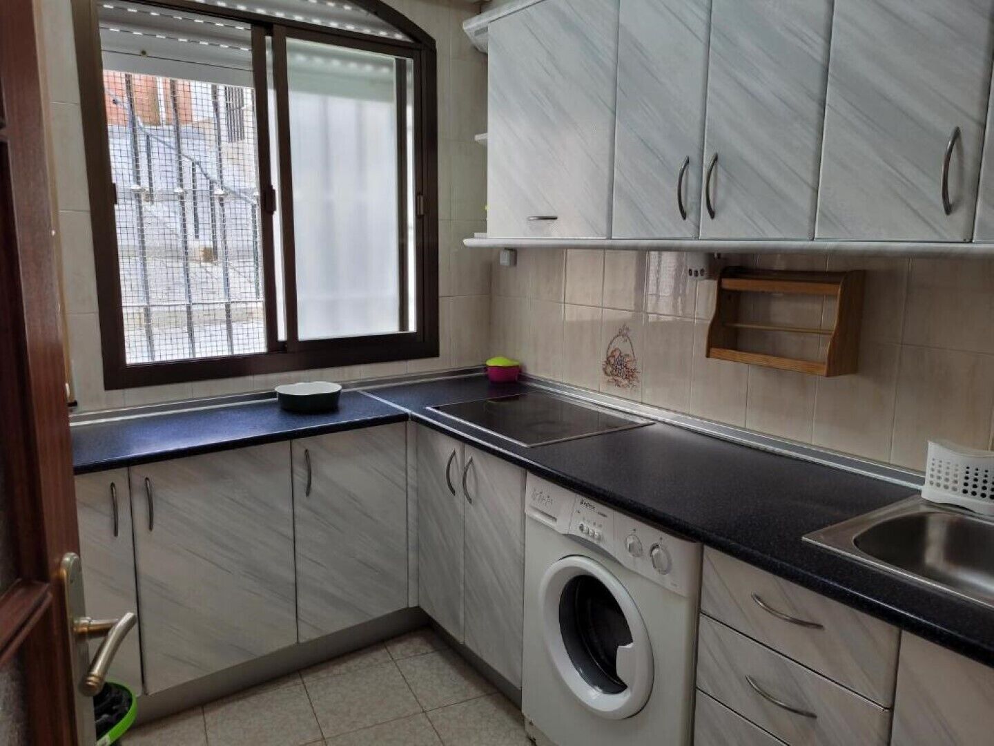 A compact kitchen with a built-in washing machine, a window with blinds, and a sink.