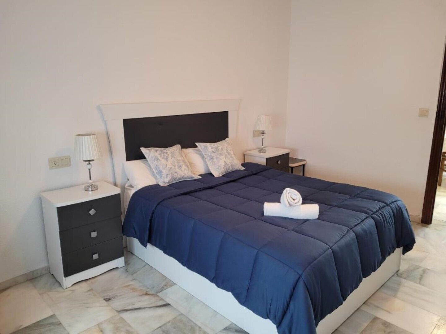 A neatly made bed with a dark blue bedspread and decorative pillows, a white bedside table with a lamp, and a small drawer unit.