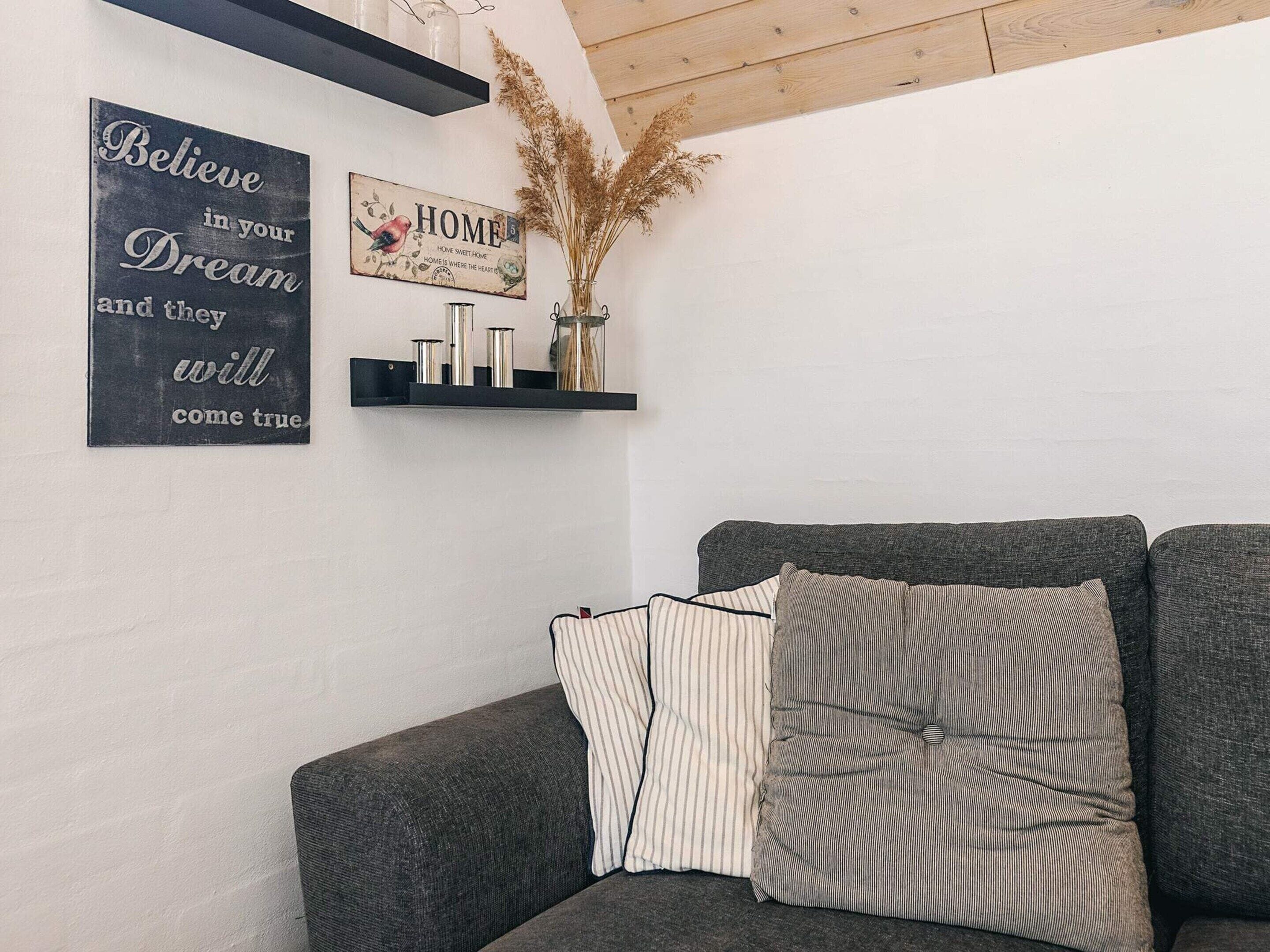 A cozy living room with a grey sofa, striped pillows, a black sign with a bird illustration, and a shelf with decorative items.