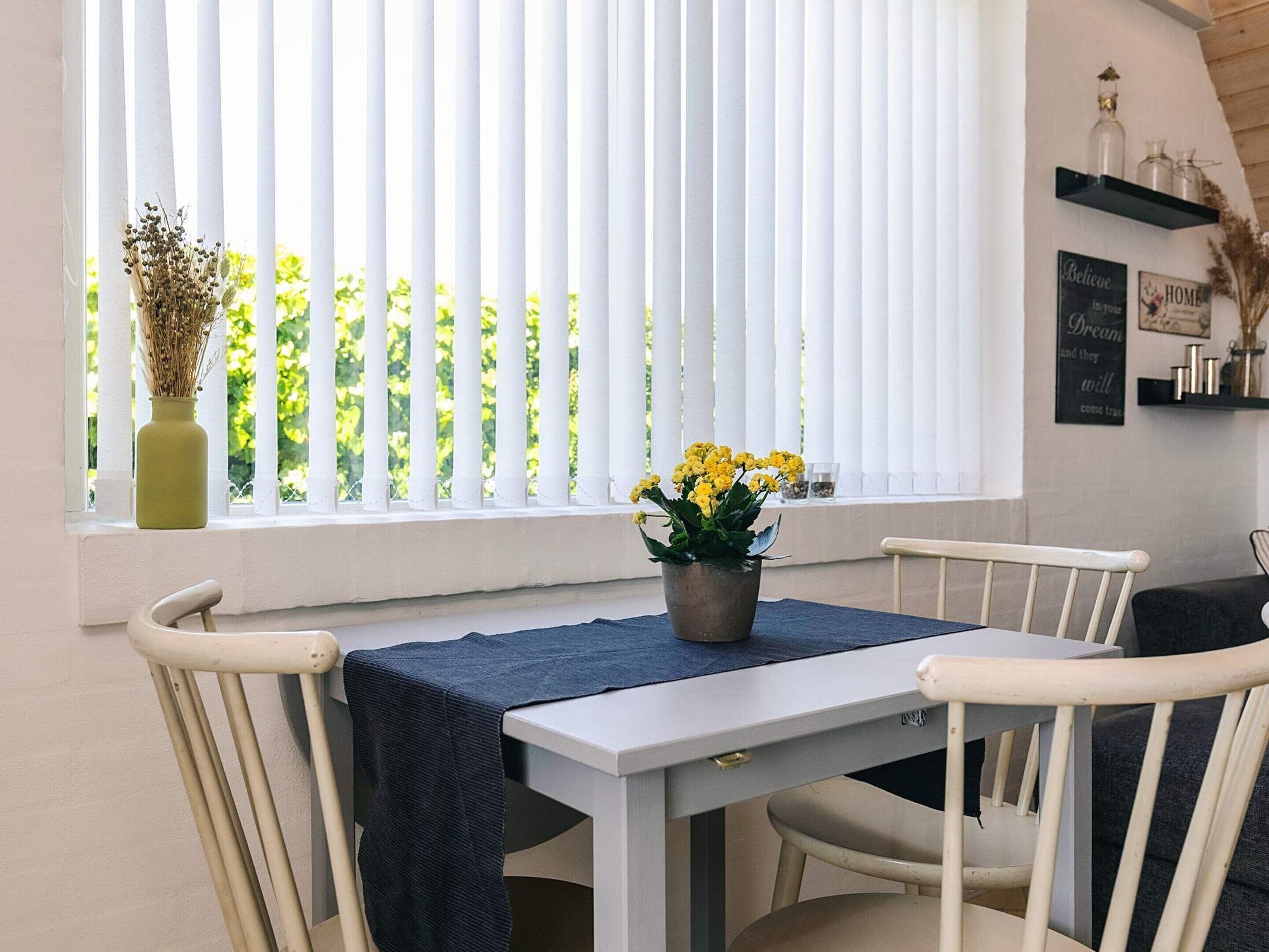 A dining area with a table set for two, white chairs, a window with vertical blinds, and a vase with yellow flowers on the windowsill.