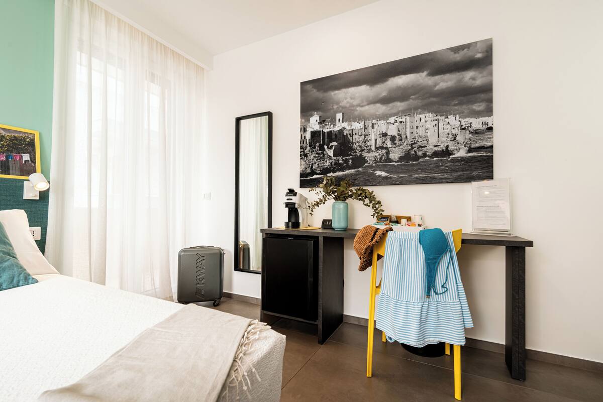 Een hotelkamer met een bed, een bureau met een computer, een stoel, een kleine tafel, een spiegel en een groot zwart-wit stadslandschap op de muur.