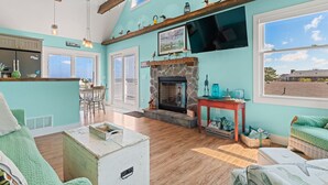 A cozy living room with a stone fireplace, a dining area with a bar, and large windows offering a view of the outside. - Ohana 3 Bedroom Home in Walnut Island (Grandy)