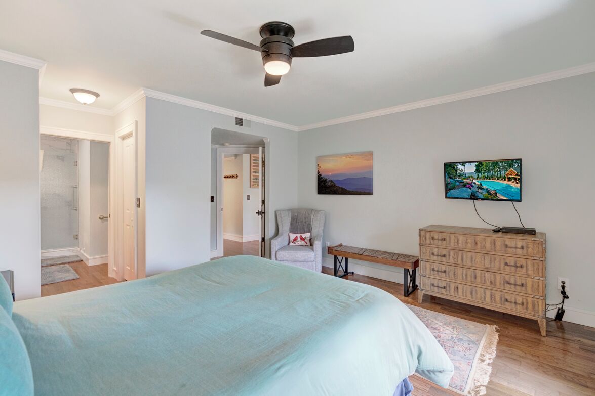 Un dormitorio con cama, ventilador de techo, televisor, cómoda de madera y una silla.