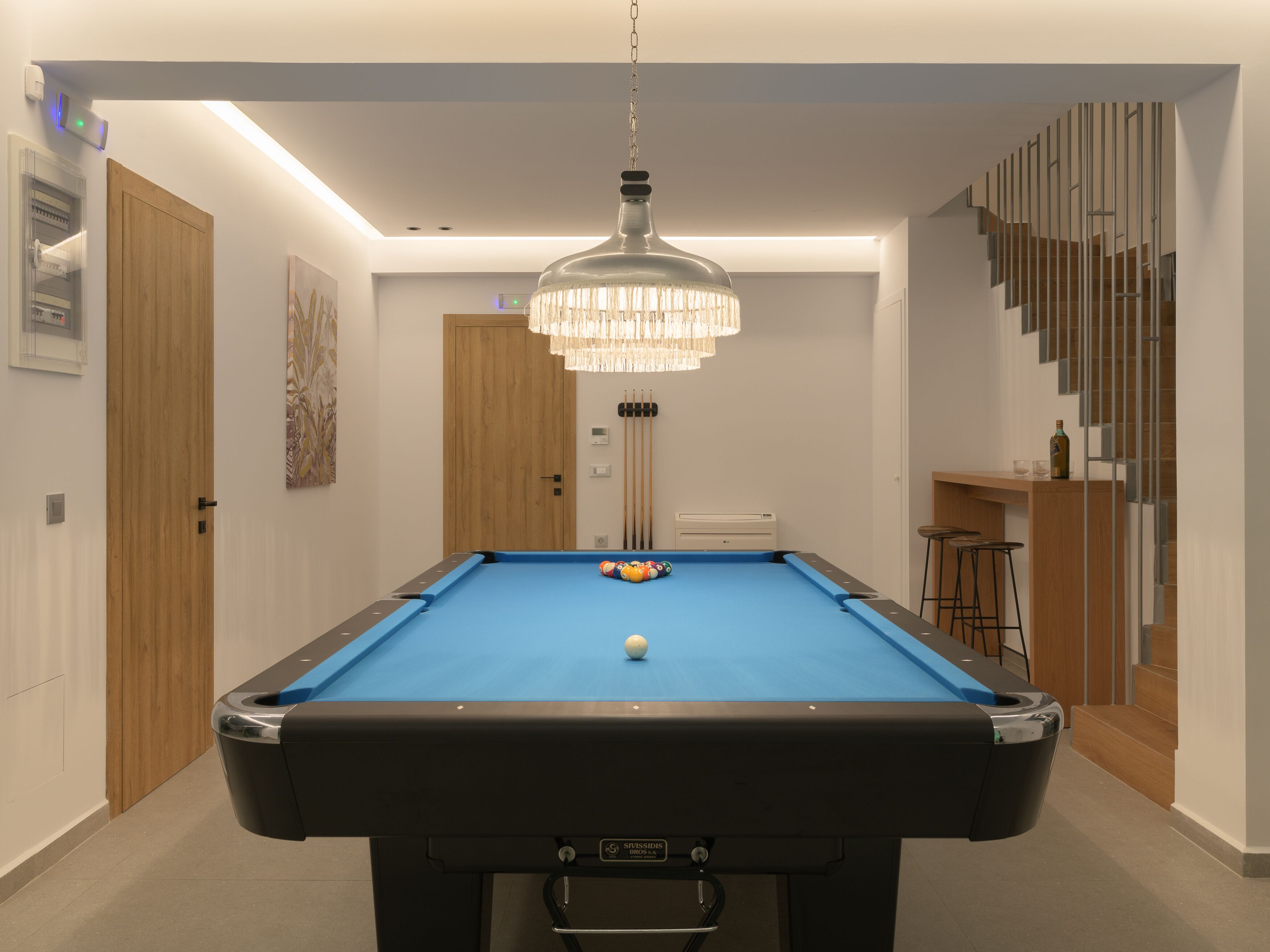 Ein Billardtisch mit blauer Spielvliesbahn, eine darüber hängende Chandelierleuchtre und eine Treppe rechts.