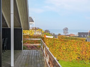 A wooden deck with a railing, a view of the sea, and a hedge with yellow leaves. - 6 Person Holiday Home in Borkop (Borkop)