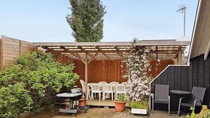 En terrasse med et træbeklædt pergola, hvide plastikstole, et bord og krukker med planter.