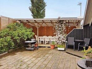 A patio with a wooden pergola, white plastic chairs, a table, and a grill.