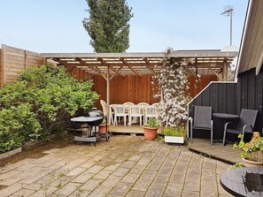 Une terrasse dotée d’une pergola en bois, de chaises en plastique blanches, d’une table et d’un barbecue.
