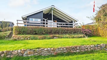 Una casa moderna con techo inclinado, un muro de piedra en primer plano y una bandera en un asta a la derecha.