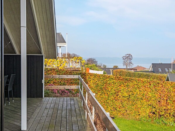 A wooden deck with a railing, a view of the sea, and a hedge with yellow leaves.