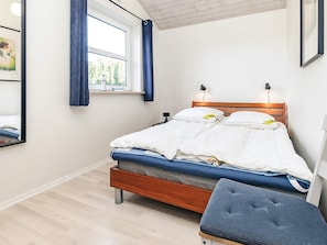 A bedroom with a wooden bed, white bedding, a window with blue curtains, and a bedside table with a lamp.