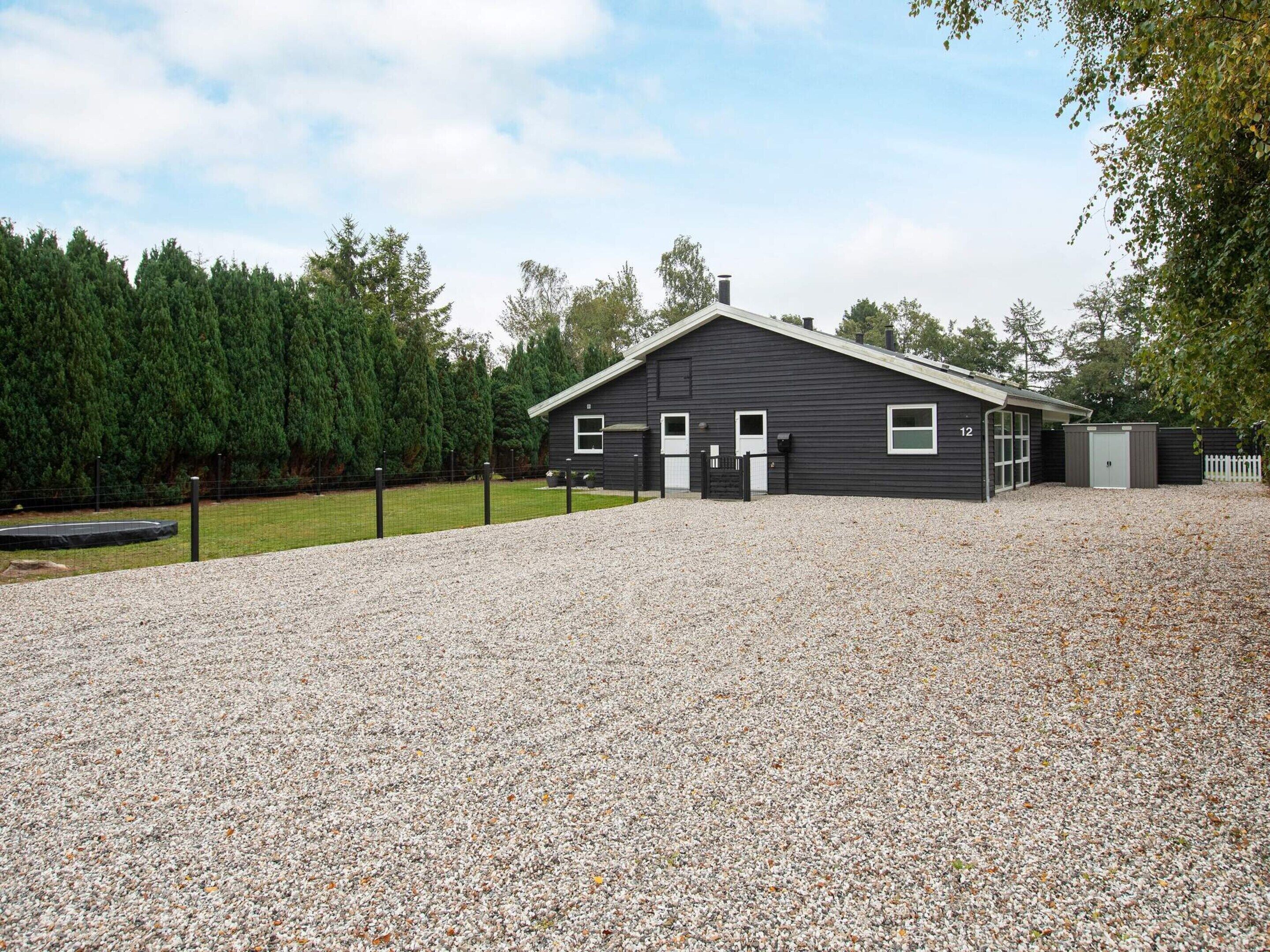 A black building with a gravel driveway, a fenced yard, and a wooded area in the background.