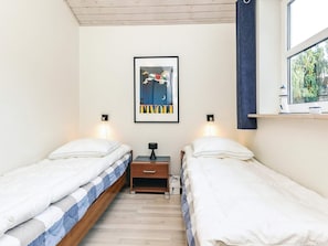 A bedroom with two single beds, a framed picture on the wall, bedside lamps, a small table, and a window with a view of trees.