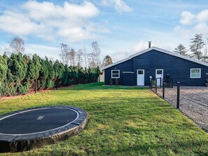 A house with a dark exterior, a front yard with a trampoline, and a row of evergreen trees.