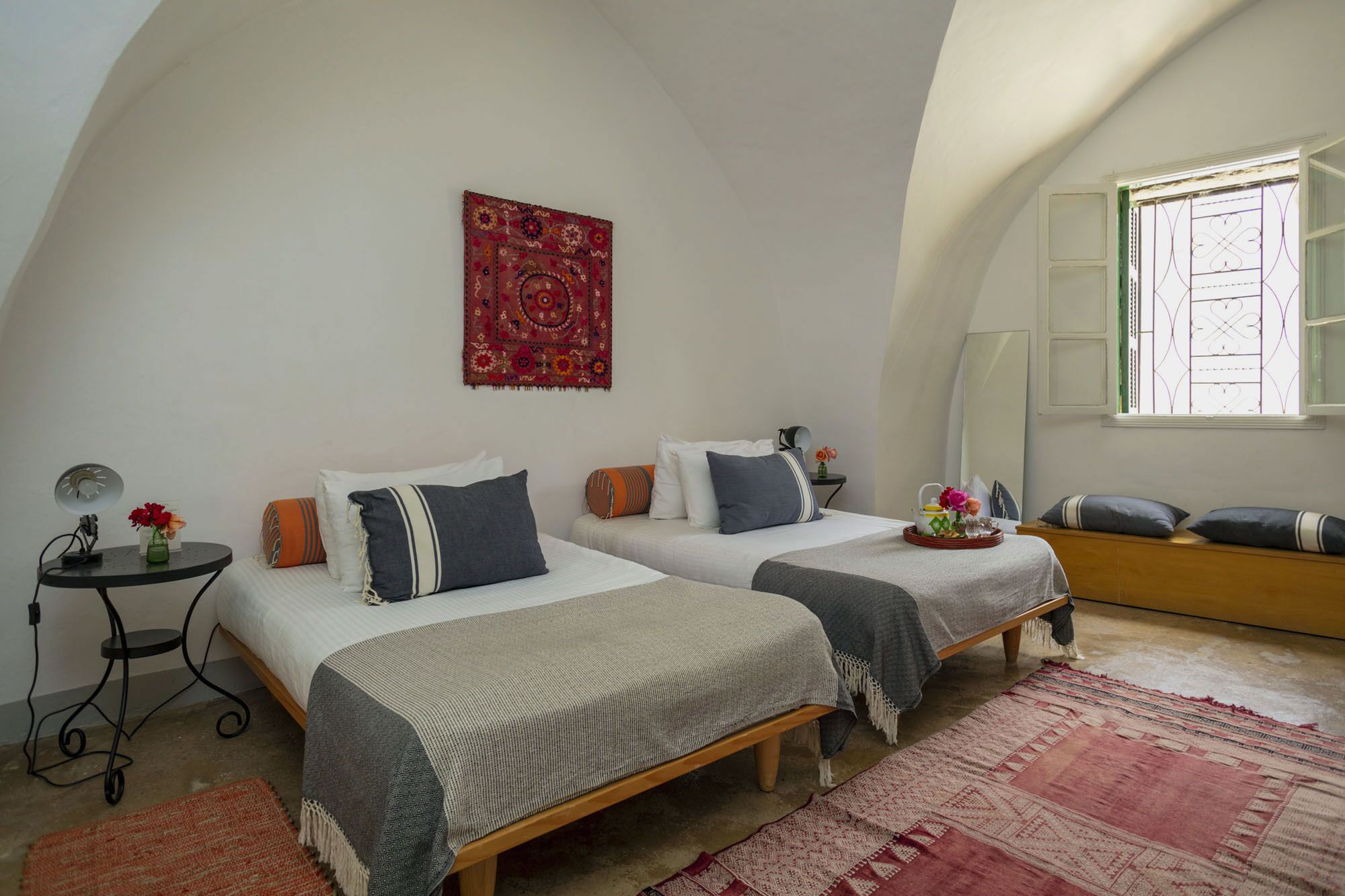 A bedroom with two beds, a red patterned tapestry, a small table with a lamp, and a window with a decorative grille.
